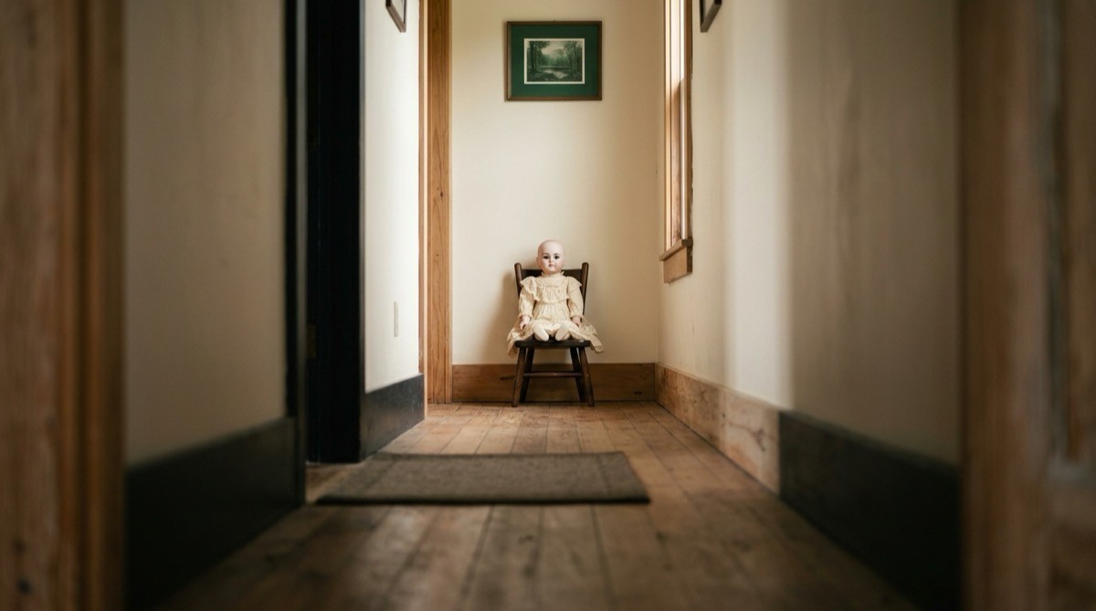 Old porcelain doll in a lace dress sitting alone in a small wooden chair in a hallway