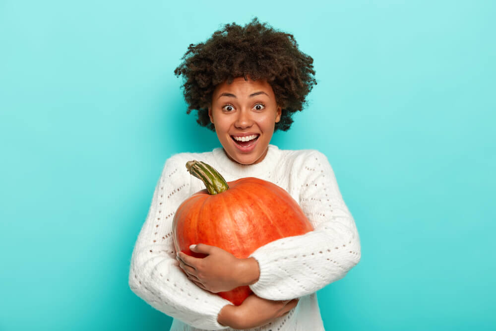 A woman who’s excited for fall wears a cozy sweater while holding a large pumpkin.