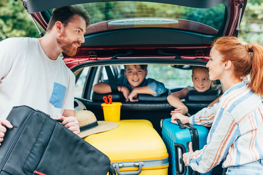 A small family packs their bags into the trunk of their car for a summer road trip.