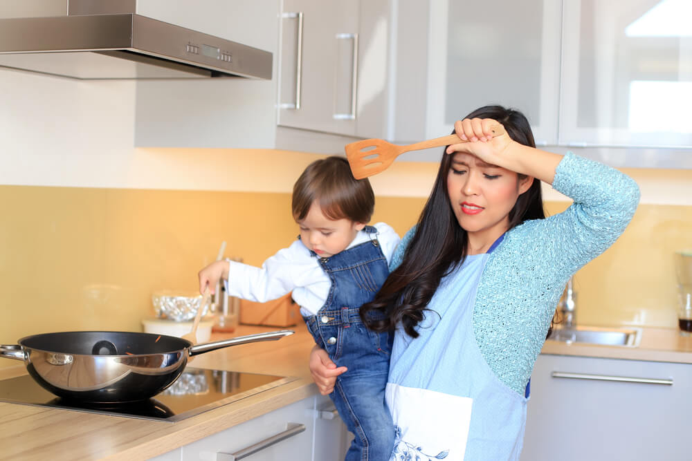 A stressed mom tries to cook dinner with her baby in one arm.