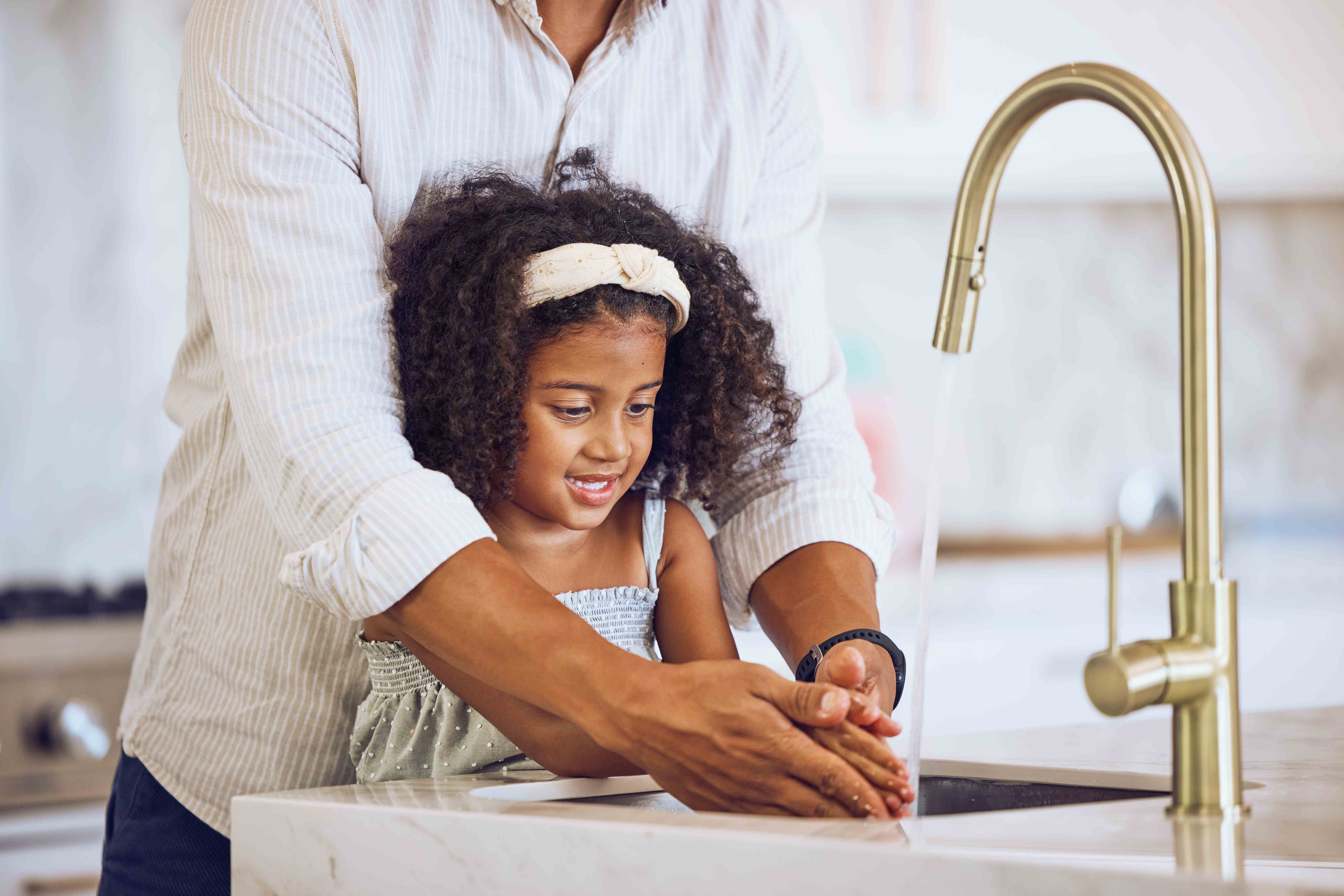A loving parent helps their daughter wash her hands in the kitchen sink