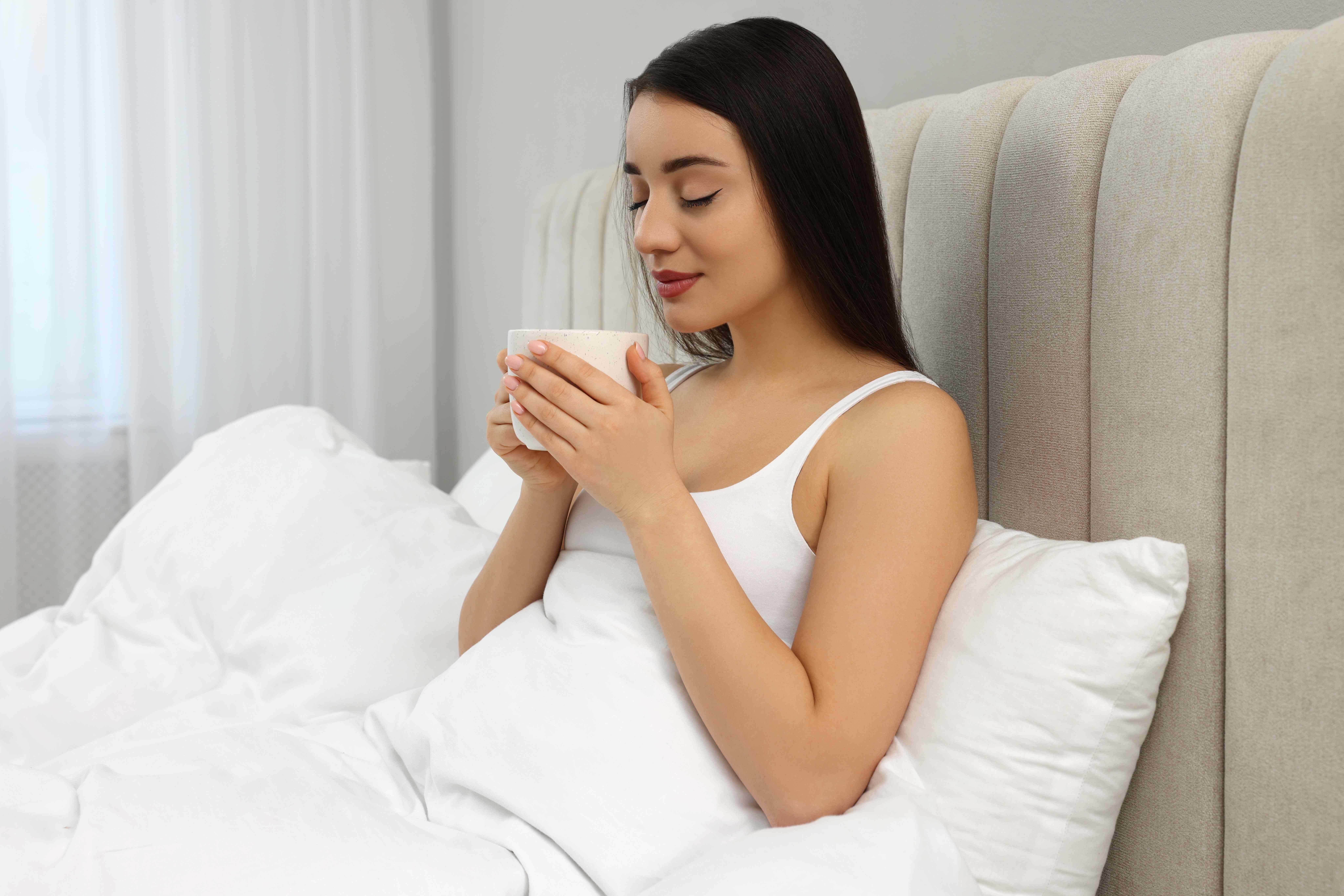  A woman relaxes in bed with a cup of herbal tea. 